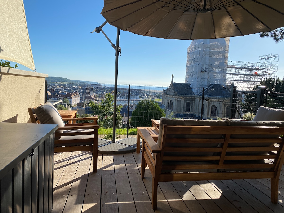 An outdoor terrace features comfortable wooden seating under a large umbrella, offering a view of the surrounding area. The horizon showcases a scenic landscape with a visible structure in the background, partially covered in scaffolding, indicating ongoing renovations.