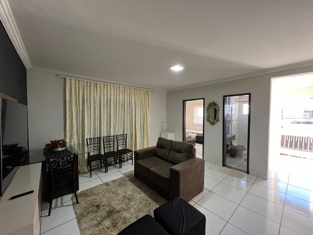 A spacious living area is displayed, featuring a cozy brown sofa and a dark chair. A light beige rug covers the tiled floor, while a rectangular table with four black chairs is positioned nearby. Soft curtains frame the large window, allowing natural light to fill the space.