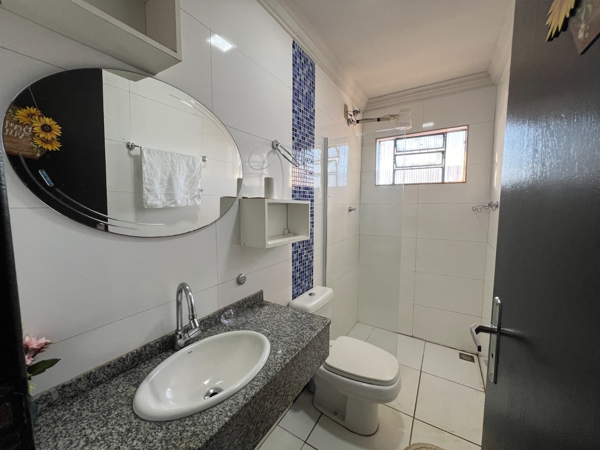A well-lit bathroom features modern fixtures and a granite sink counter. An oval mirror reflects the space, while a white towel is neatly hung on the wall. A window provides natural light, and a small shower area is visible in the corner.