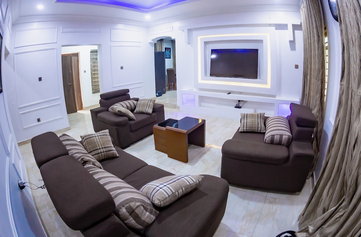 A contemporary living area is showcased with a dark sectional sofa, accented by throw pillows in various patterns. A sleek flat-screen TV is mounted on the wall, and a wooden coffee table sits in the center. Soft lighting creates a relaxed ambiance throughout the space.