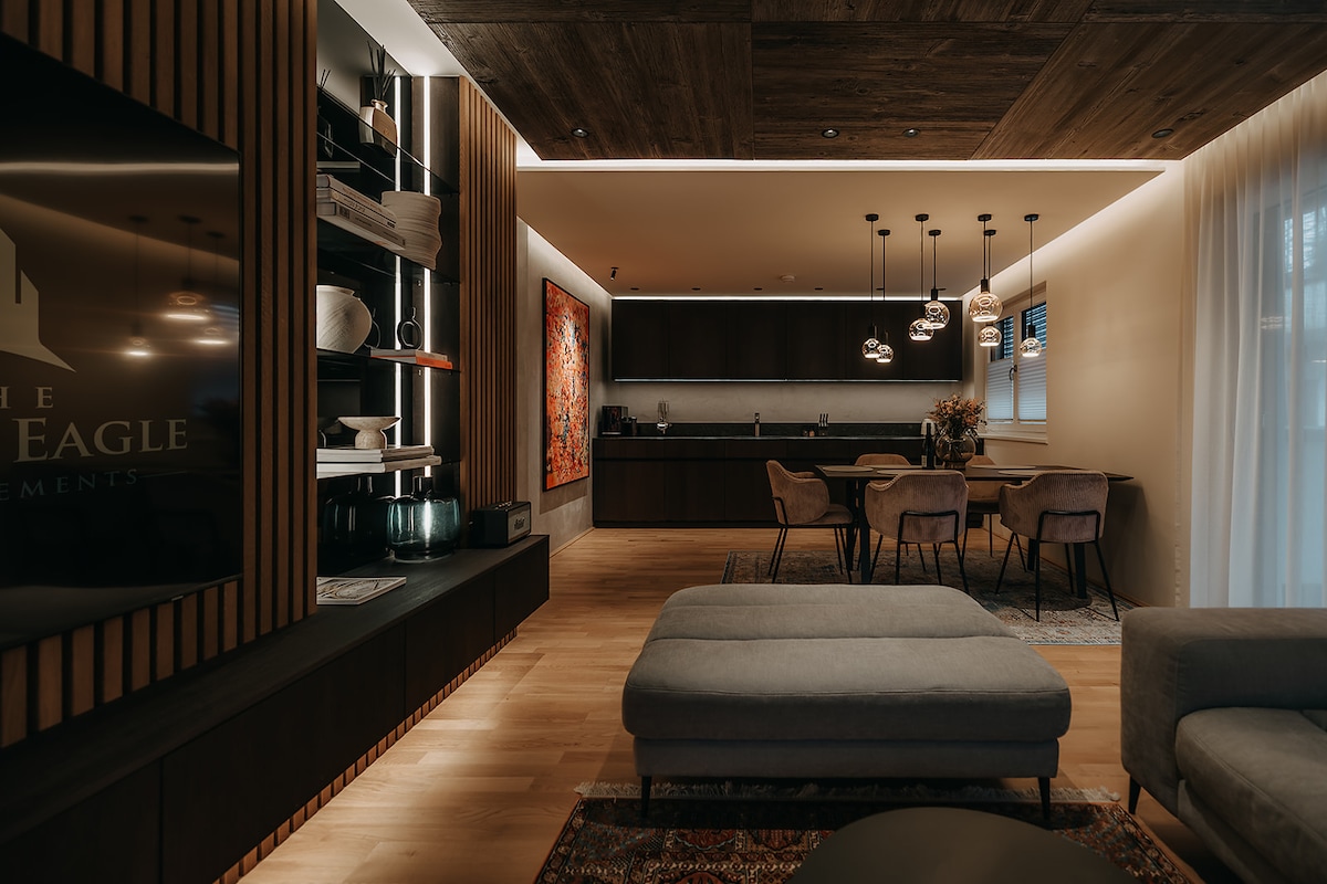 A modern living area is presented with wood paneling and elegant lighting. The kitchen features dark cabinets and a dining table surrounded by upholstered chairs. A comfortable couch is positioned in the foreground. Artwork and decorative items add visual interest to the space.