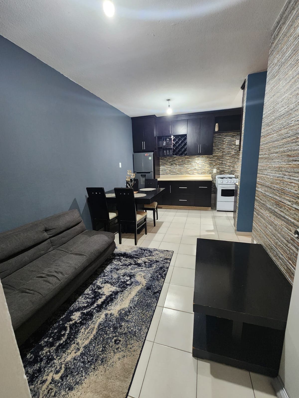 A modern living space is featured, with a dark sofa and dining table set for four. The kitchen area includes dark cabinetry and stainless-steel appliances. A light-colored wall contrasts against the deep blue and patterned accent, creating a balanced ambiance.