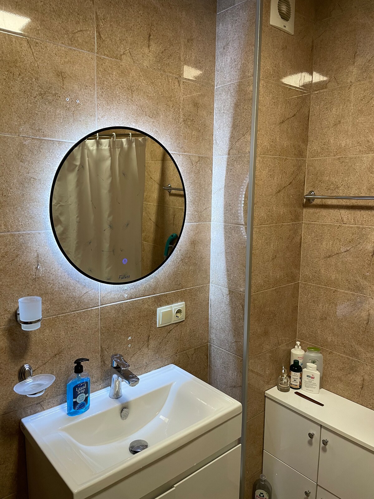 A modern bathroom features warm beige wall tiles that reflect light. A circular mirror with built-in lighting is positioned above a sleek vanity with a single sink. Essential toiletries are organized on the counter, and a shower area is visible in the background.