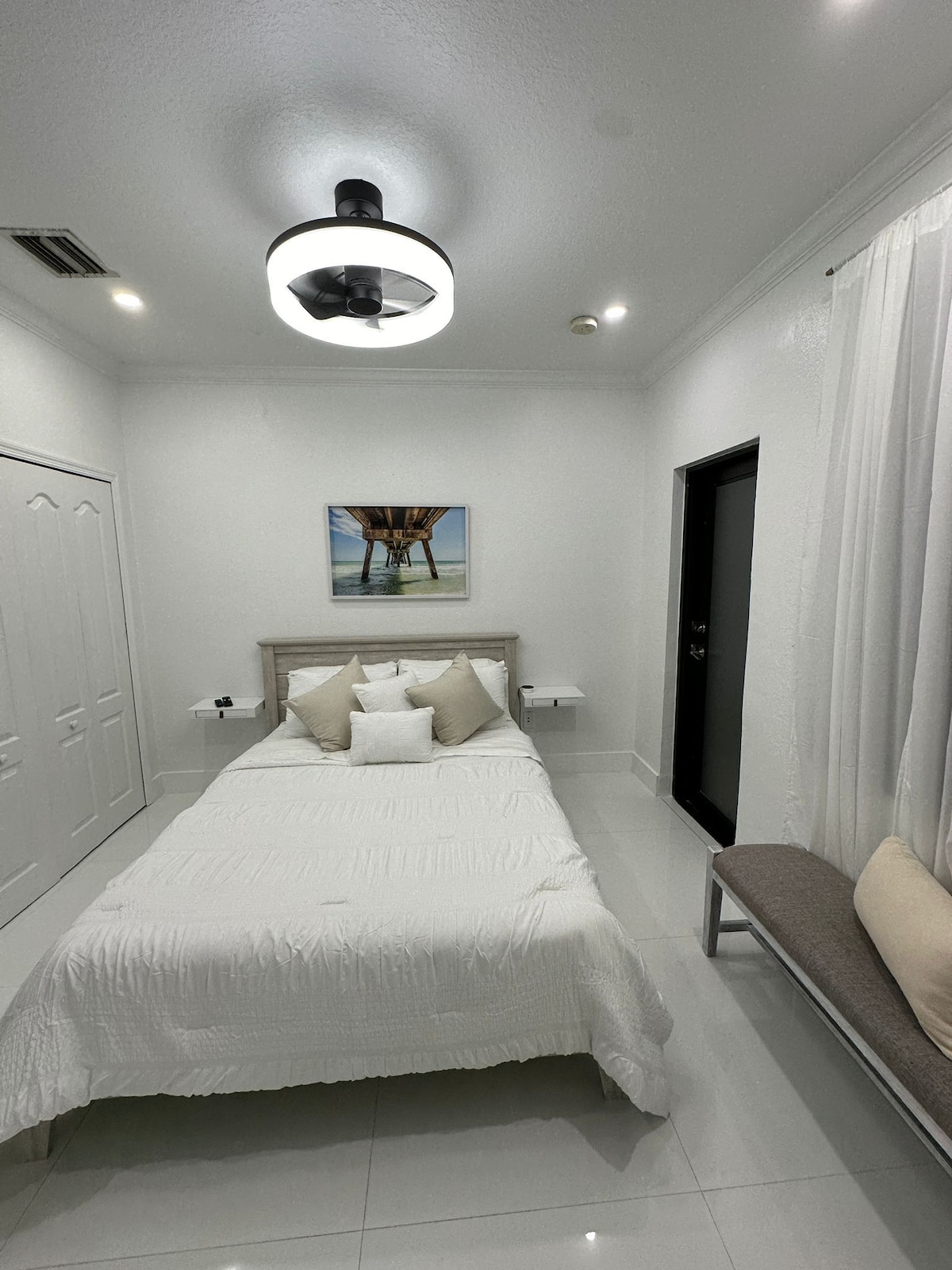 A bright sleeping area features a spacious bed adorned with a white quilt and multiple decorative pillows. A piece of wall art depicting a pier is displayed above the bed. Nightstands are positioned on either side, and natural light enters through a window with sheer curtains.