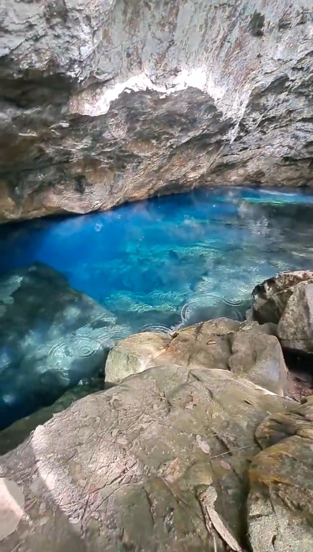 A serene natural pool is surrounded by rugged rock formations, with crystal-clear blue water reflecting the light. The texture of the rocks and the gentle ripples on the water's surface create a peaceful scene, inviting exploration in a tranquil setting.