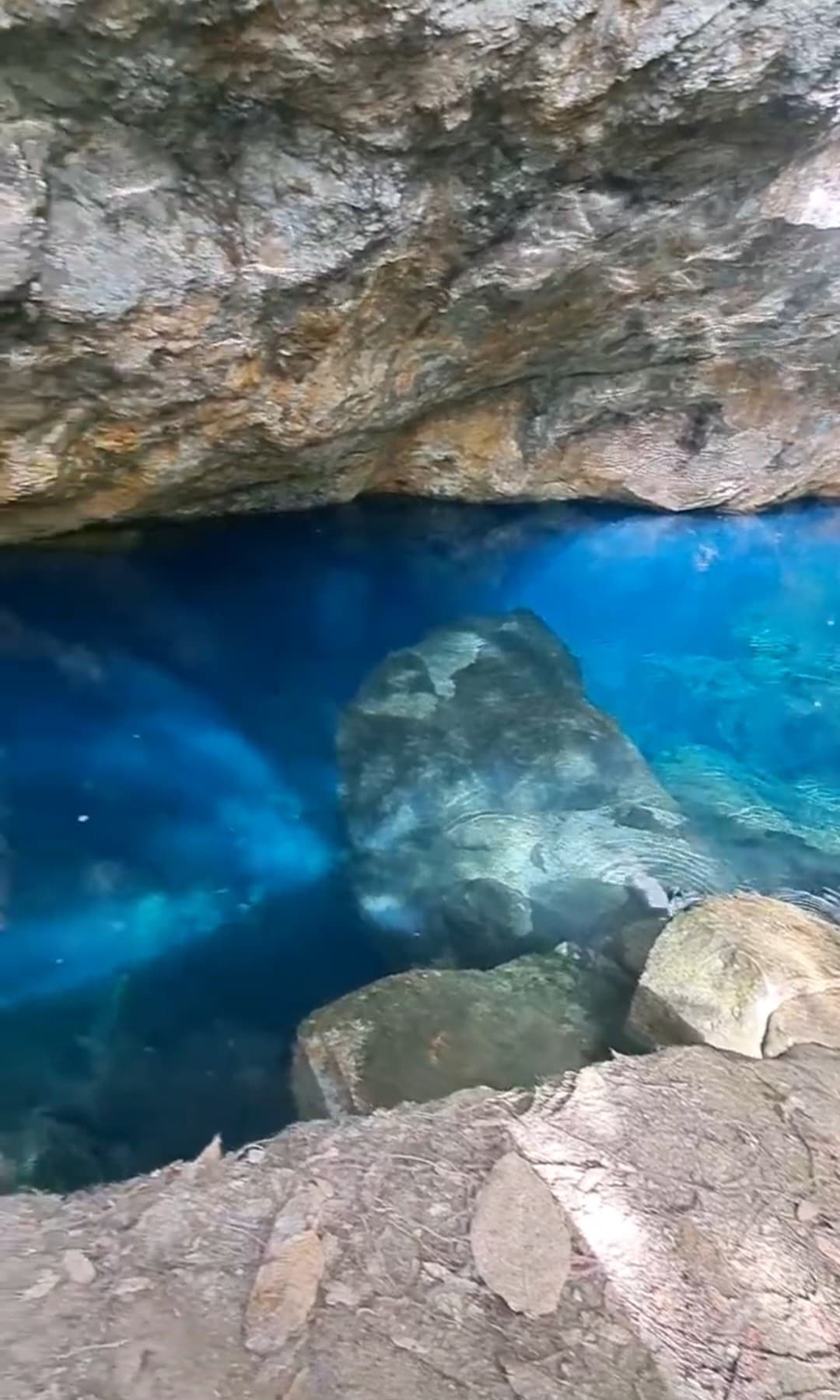 A serene natural pool is framed by rugged stone walls, featuring clear blue water that reveals underwater textures. Smooth rocks and pebbles line the water's edge, and light reflects gently off the surface, creating a tranquil atmosphere.