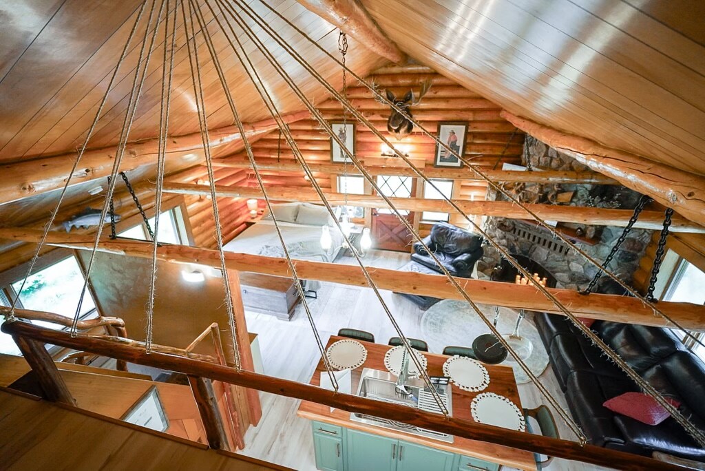 An aerial view of the open concept studio cabin reveals wooden beams and a high ceiling. A well-equipped kitchen with plates arranged on the counters is visible, alongside a cozy seating area featuring dark furniture. Natural light streams through large windows, illuminating the space.