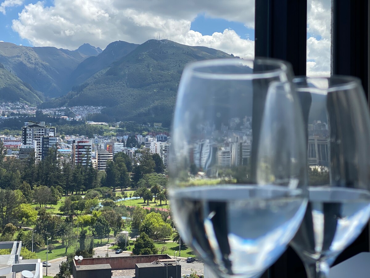 Two wine glasses filled with water are positioned in the foreground, set against a panoramic view of Quito's landscape. Lush greenery and city buildings are visible beneath a backdrop of mountains and a partly cloudy sky.