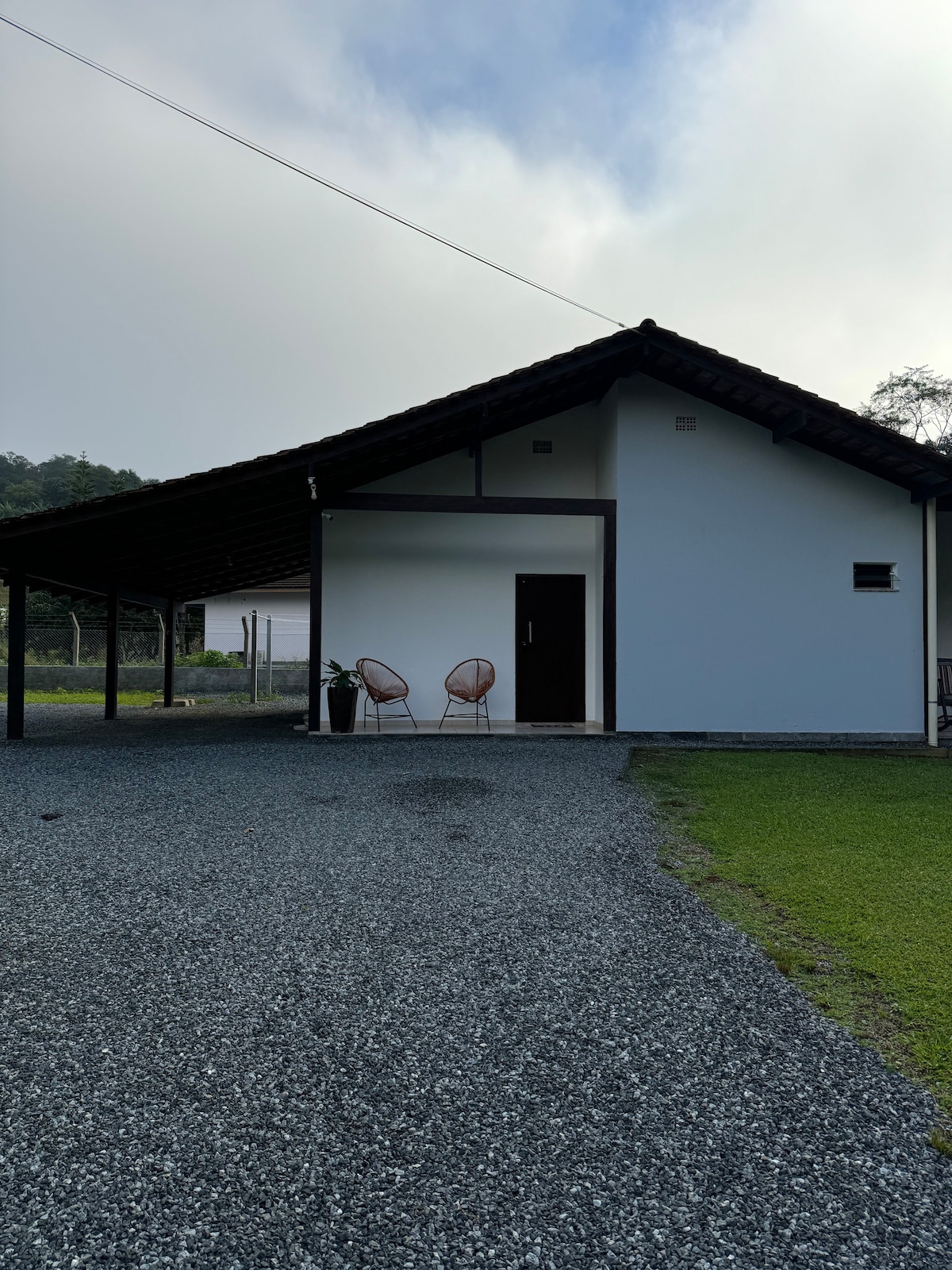 A single-story house with a sloped roof is set against a backdrop of greenery. Two wicker chairs are positioned on a gravel area near the entrance, providing a space for relaxation. A neatly trimmed lawn and natural surroundings are visible in the background.