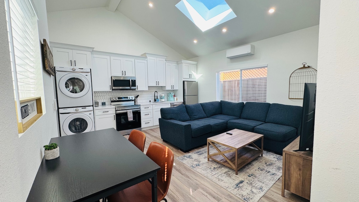 An open living space is presented, showcasing a large sectional sofa facing a coffee table. A modern kitchen is equipped with stainless steel appliances, including a washer and dryer. Natural light pours in through a skylight, enhancing the airy ambiance.