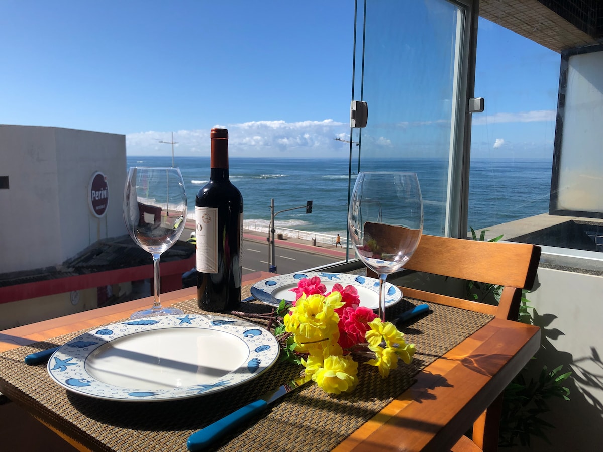 A dining table is set for two, featuring a dark bottle of wine and two clear wine glasses. Colorful artificial flowers are arranged in the center of the table. The backdrop includes a panoramic view of the ocean and a clear blue sky.