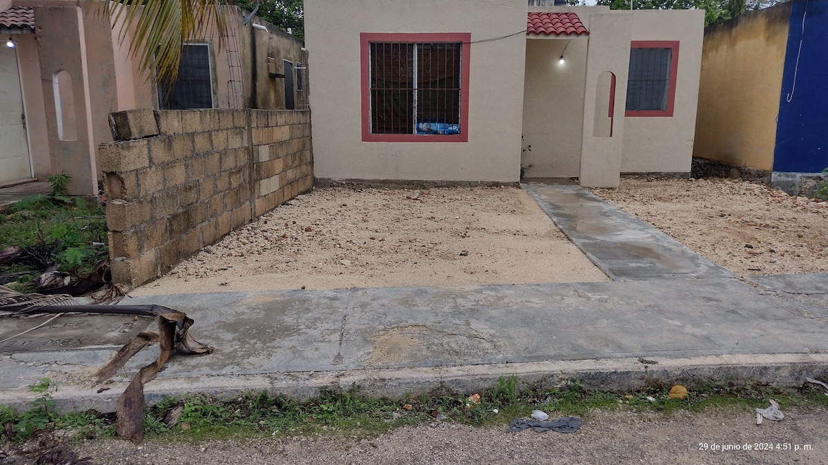 The exterior view of a single-story house with a light-colored facade and a red-tiled roof is shown. A small path leads to the entrance, with an unfinished yard made of compacted earth and a low stone wall visible to the side.