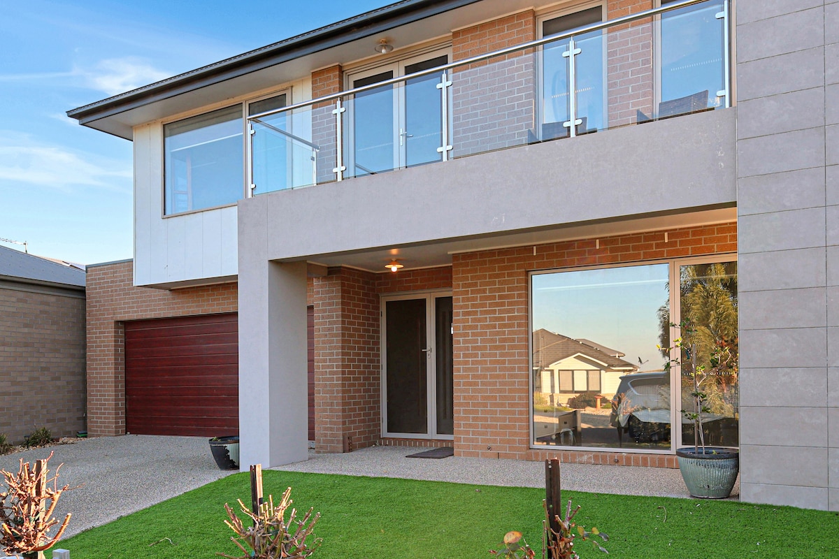 The exterior of a modern two-story home is showcased, featuring a blend of brick and smooth paneling. A balcony with glass railings is visible on the upper floor, while a doorway leads to the entrance. Well-maintained greenery frames the entrance, providing a welcoming appearance.