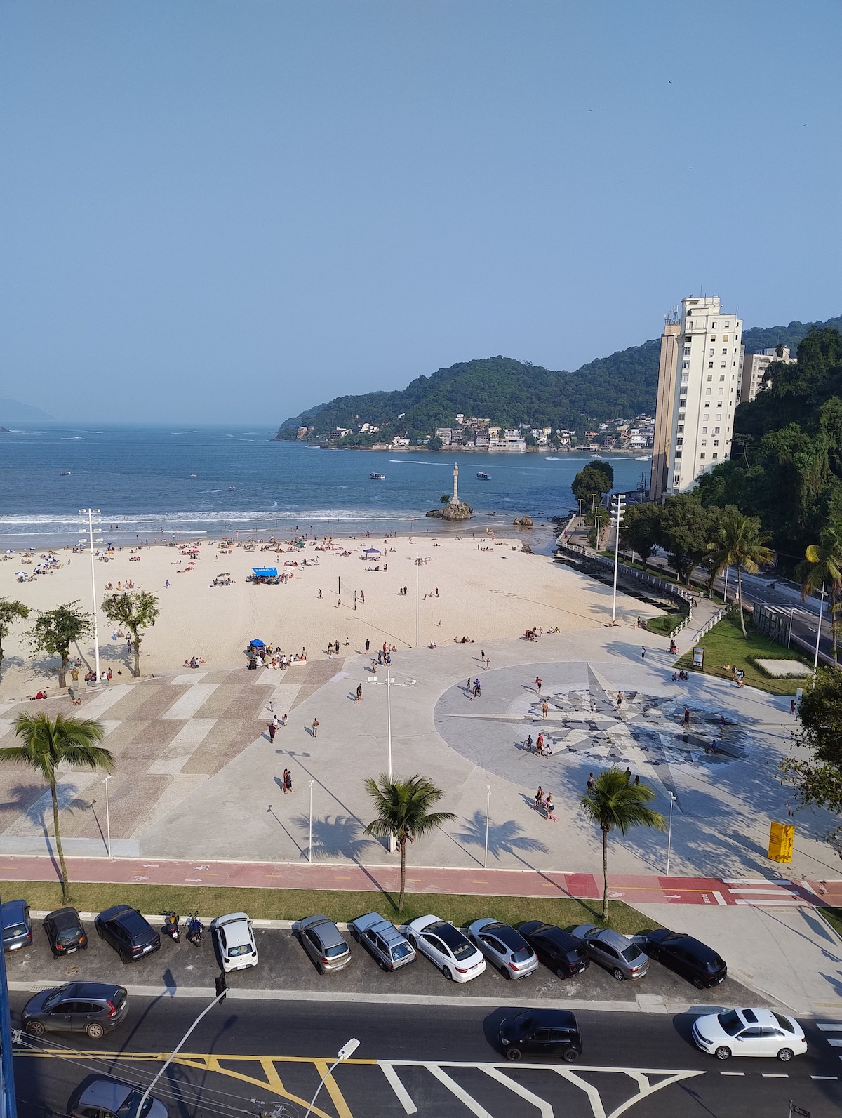 A view captures the sandy beach and coastal landscape, with visitors enjoying the area. The ocean reflects blue skies, while palm trees line the promenade. Vehicles are parked along the street, and a modern building stands in the background, completing the scenic environment.