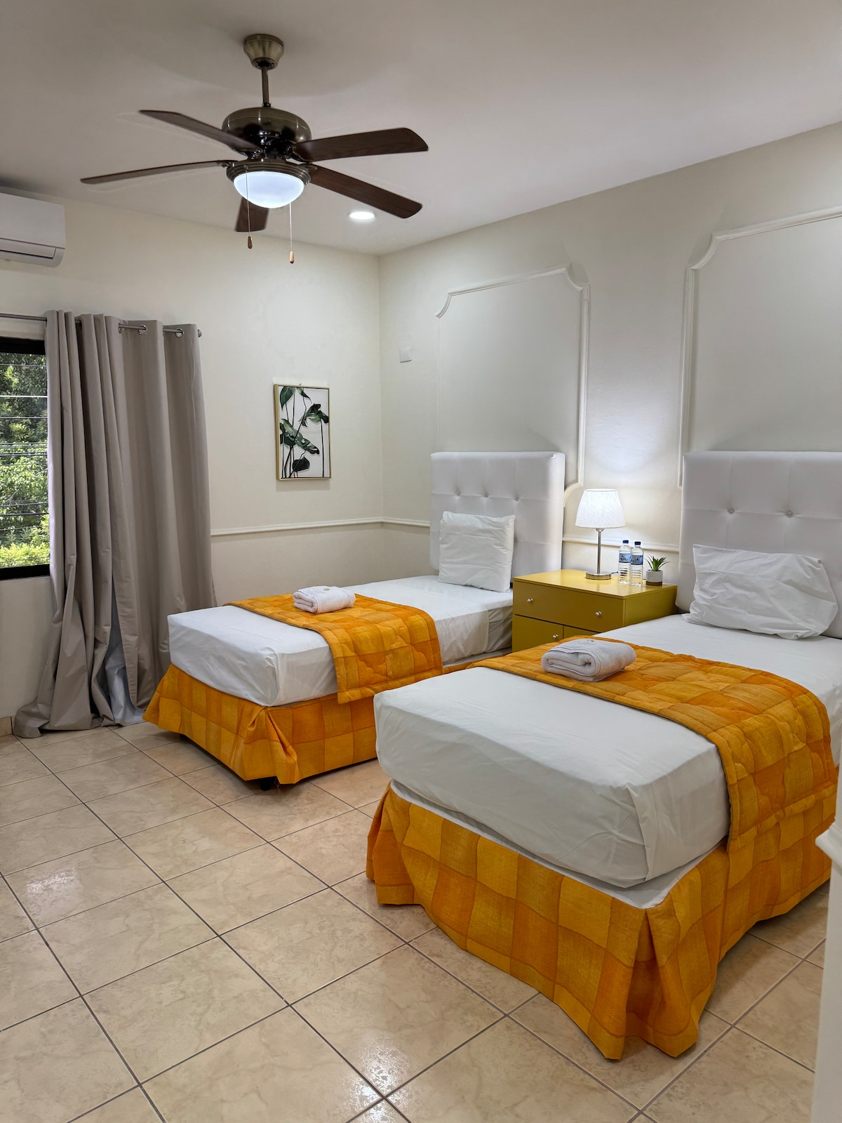 Two twin beds are situated in a bright room accented with white walls and a fan overhead. Each bed is dressed in white linens and an orange checkered blanket, accompanied by a towel. Natural light filters through a nearby window, enhancing the spacious feel.