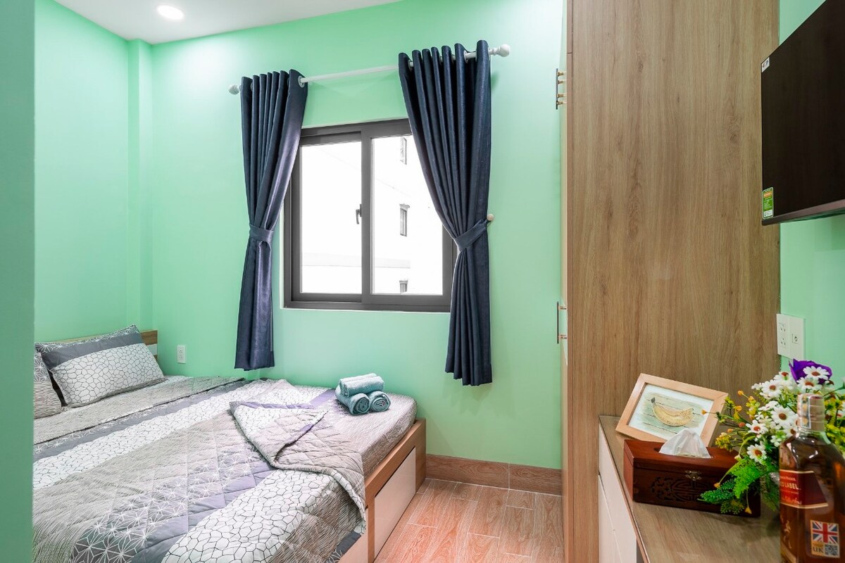 A cozy sleeping area features a spacious bed with patterned bedding. Light green walls complement dark curtains framing a window that allows natural light to enter. A wooden wardrobe is positioned nearby, and decorative elements are arranged on a dresser.