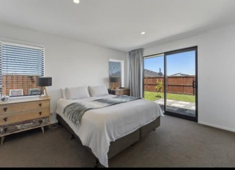 A serene bedroom features a spacious bed dressed in neutral linens, complemented by a soft throw. Natural light enters through two windows and a sliding door, which leads to an outdoor area. A stylish dresser and lamp contribute to the room's functionality.