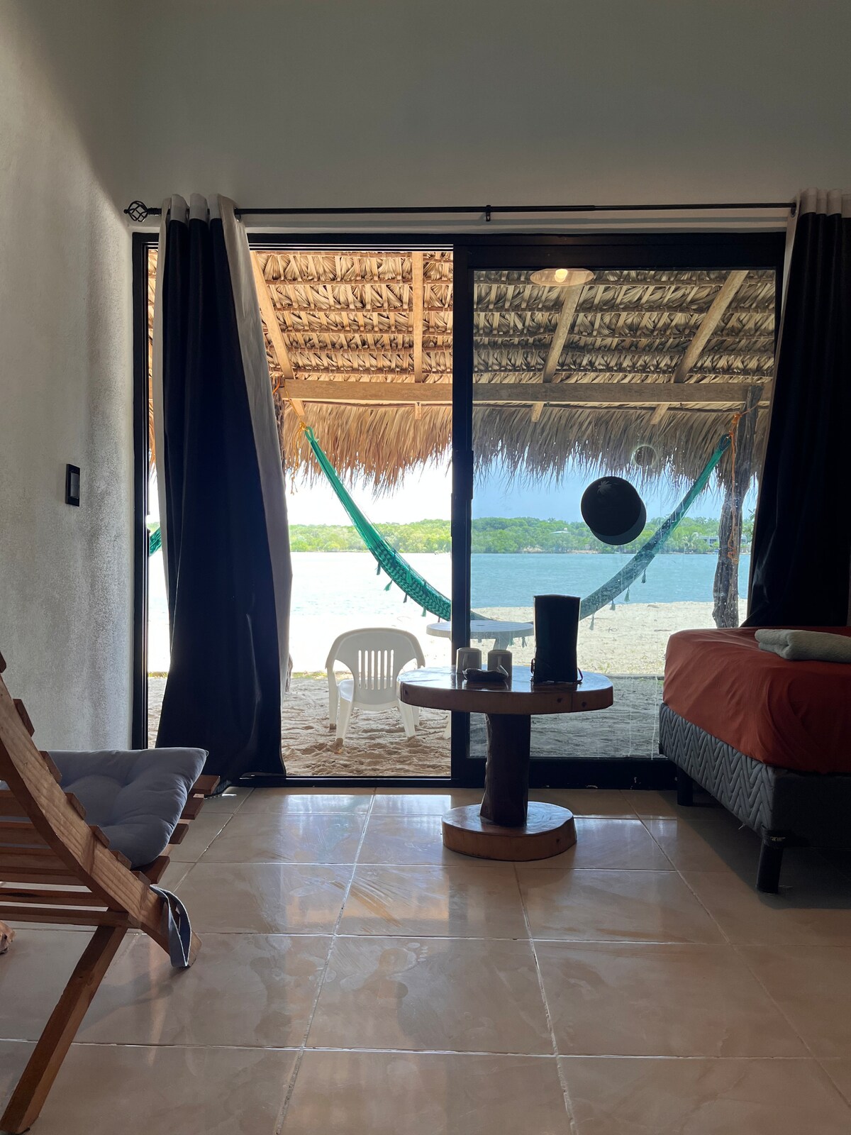 The interior space features a comfortable seating area with a wooden chair and a round table. A sliding glass door opens to a view of a sandy beach with hanging hammocks and lush greenery beyond. Natural light fills the room, complemented by dark curtains.