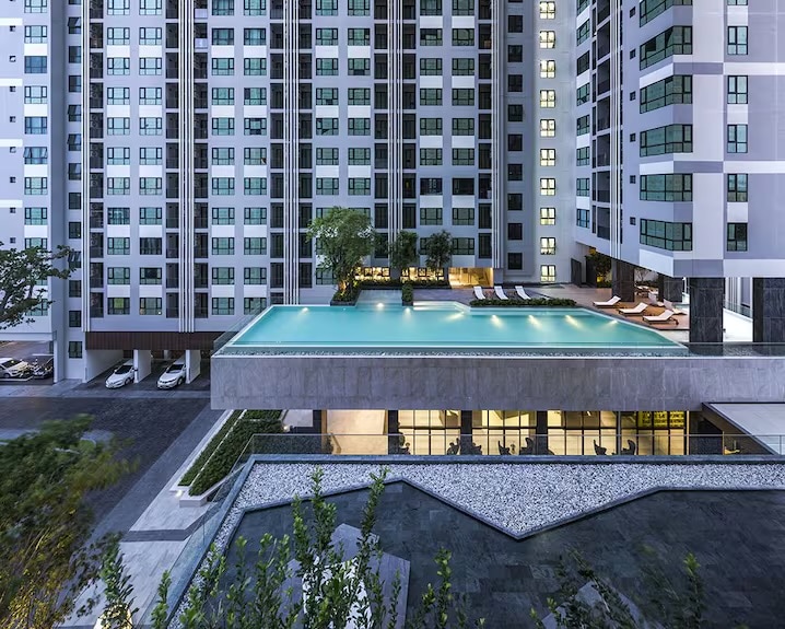 An elevated infinity pool glistens under evening lights, surrounded by a modern multi-story condo complex. Poolside loungers can be seen arranged neatly for relaxation, with greenery adding a touch of nature to the contemporary architecture.