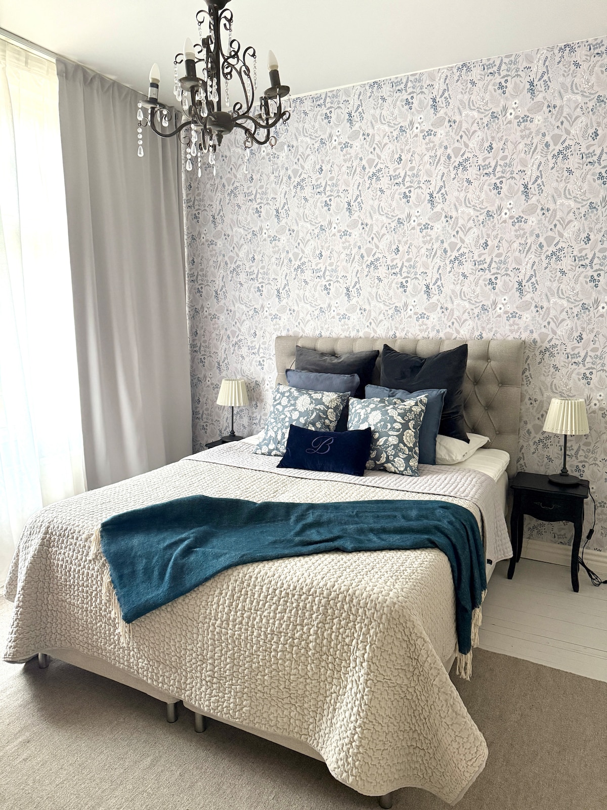 A cozy bedroom features a tufted bed with decorative pillows and a textured blanket. Soft lighting from lamps on either side complements the floral wallpaper and elegant chandelier overhead. Natural light filters through sheer curtains, enhancing the room's tranquil atmosphere.