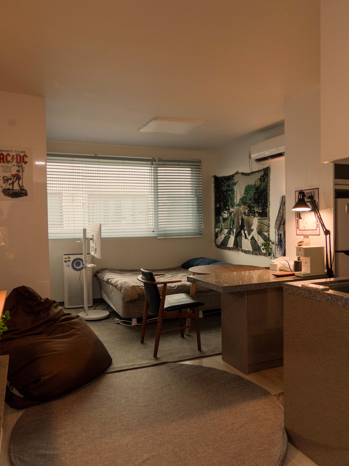 An open living space features a comfortable bed beside a large window with horizontal blinds. A small round rug lies on the floor, with a wooden table and a chair positioned nearby. A wall tapestry depicting a scenic view adds interest to the room.