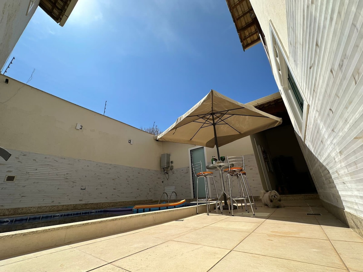 A sunny outdoor area is captured, featuring a small swimming pool and a shaded seating arrangement under a large umbrella. The spacious patio is paved with light-colored tiles, providing an inviting space for relaxation. The surroundings include smooth white walls and clear blue skies.