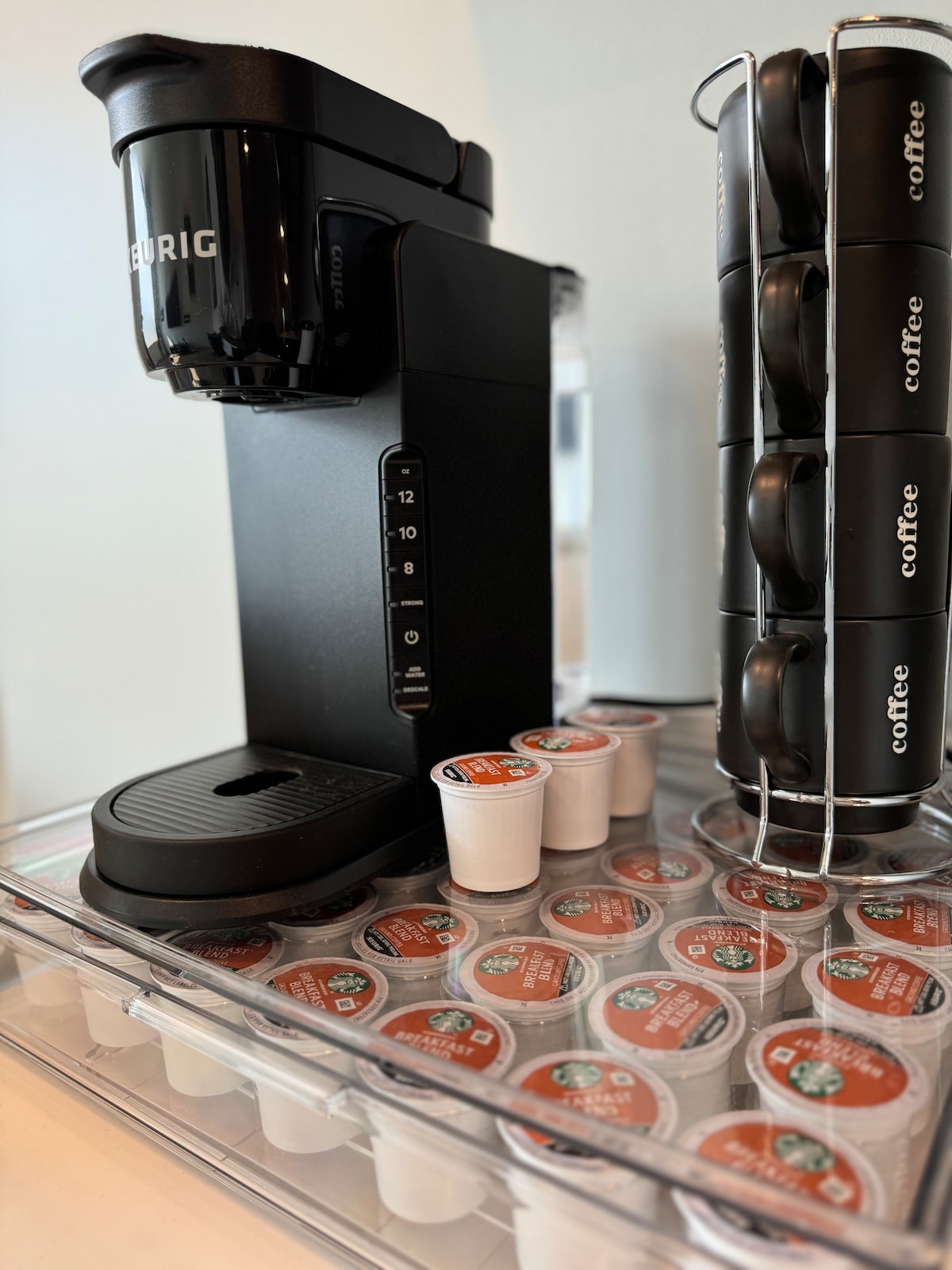 A Keurig coffee maker is positioned on a clear tray, accompanied by a selection of coffee pods. The pods are organized in a vertical holder, enhancing accessibility for guests. The setup creates a convenient coffee station for morning routines.
