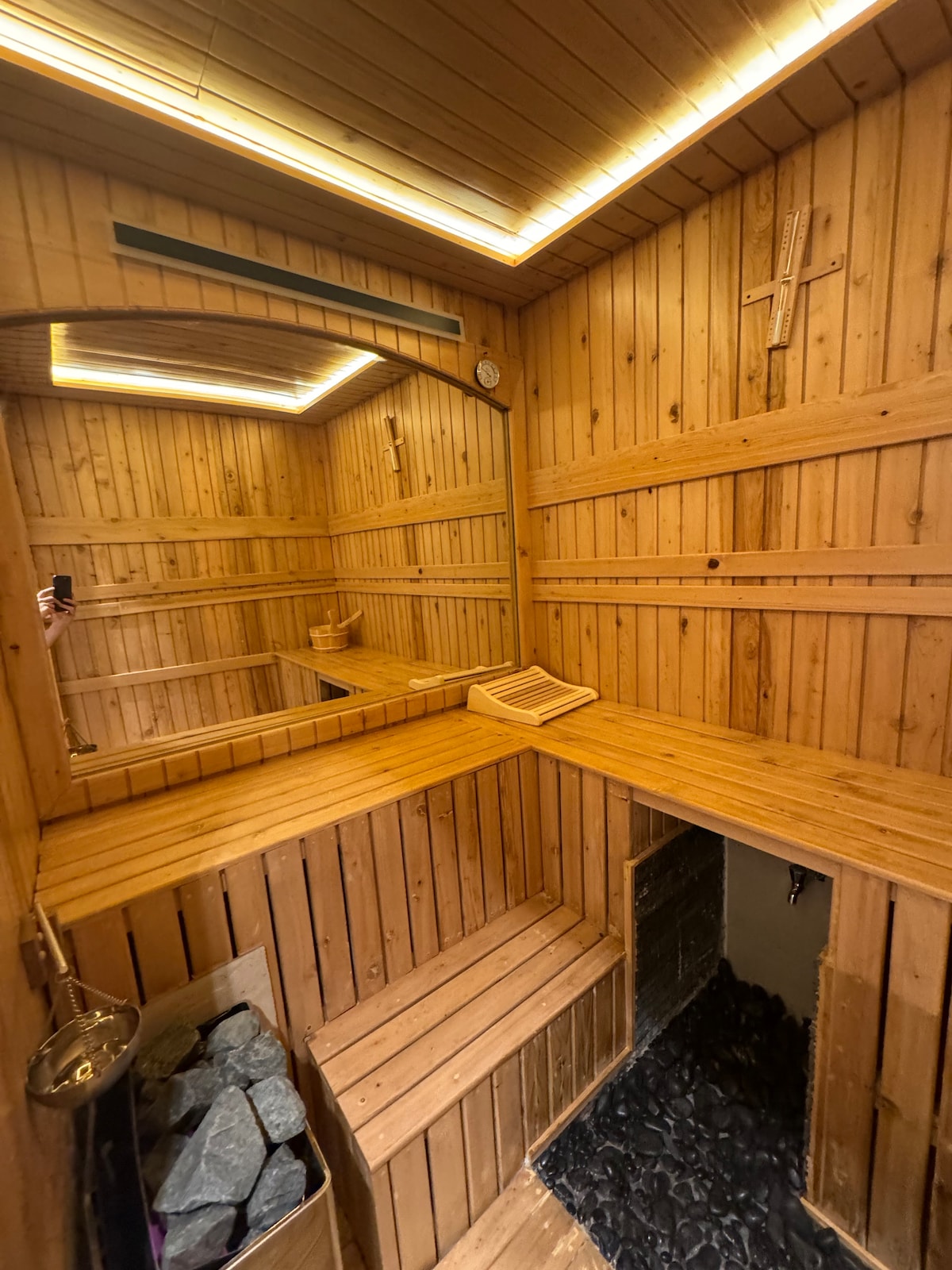 A wooden sauna features warm, natural wood paneling and ambient ceiling lighting. Benches are arranged for seating, with a stone heater visible at one side. The space includes simple decor elements such as a clock and a cross, contributing to a calming atmosphere.