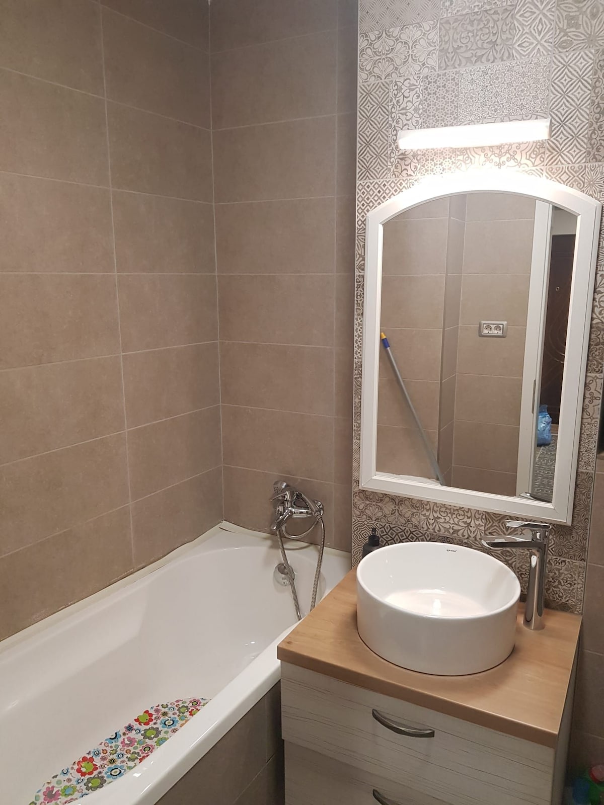 A modern bathroom features a white bathtub with a decorative floral pattern at the base. A circular sink is positioned on a wooden countertop, accompanied by a sleek faucet. A mirror above the sink enhances the sense of space, and soft lighting is provided by a mounted fixture.