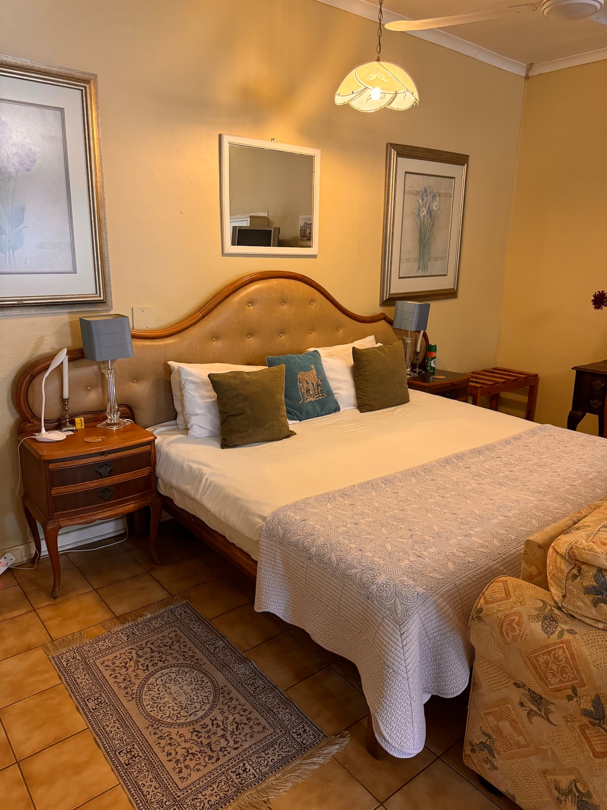 A cozy sleeping area features a spacious bed with a tufted headboard, complemented by two decorative pillows. A bedside table on each side holds lamps, and natural light filters in through a nearby window, illuminating the soft beige walls and warm tile flooring.
