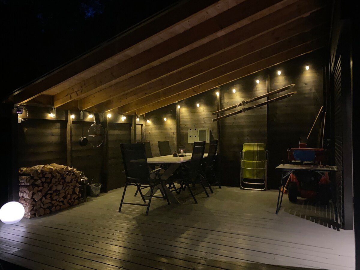 An outdoor terrace features a spacious table surrounded by six chairs, illuminated by soft lighting along the wooden beams. A stack of firewood is positioned to one side, and equipment is neatly arranged for convenience. The area is designed for relaxation and enjoyment.