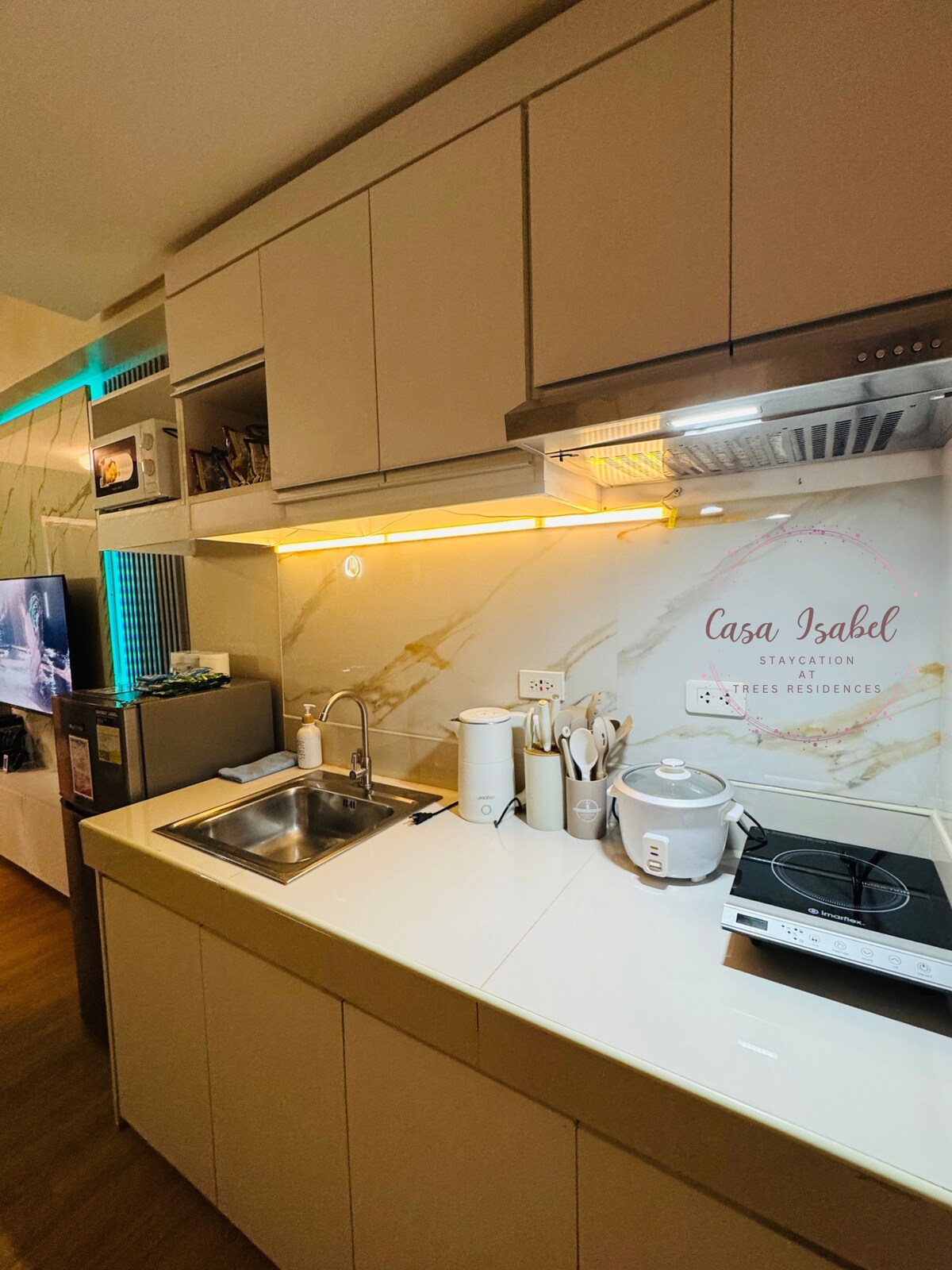 The compact kitchen area features sleek cabinetry and a marble backsplash. A double sink and stovetop are positioned on the counter, accompanied by a kettle and rice cooker. Soft lighting enhances the workspace, while modern appliances provide convenience.