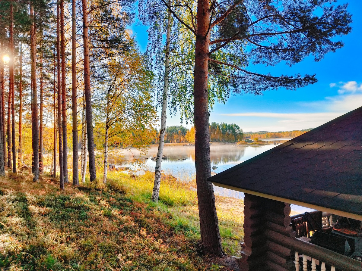 Tall trees frame a serene lakeside view, with calm waters reflecting the clear sky. The landscape is adorned with autumn foliage, creating a peaceful natural setting. A cabin roof is visible in the foreground, suggesting proximity to this tranquil environment.