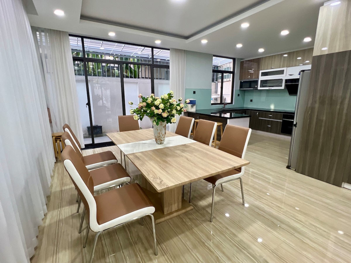 A dining space featuring a large wooden table surrounded by six modern chairs. A vase of fresh flowers sits at the center. Bright lights illuminate the area, highlighting the sleek cabinetry and spacious kitchen visible in the background through large glass doors.
