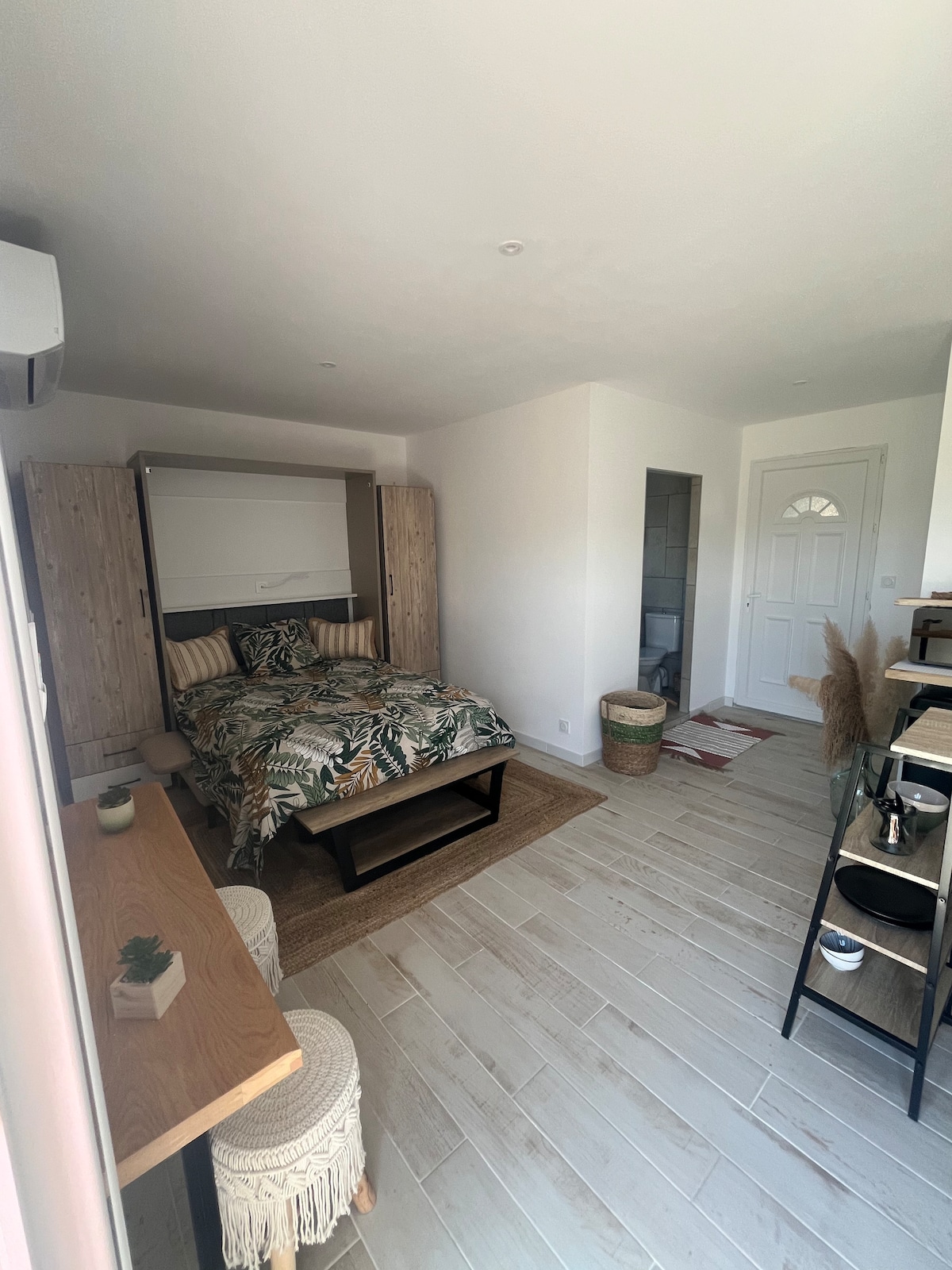 A cozy studio is presented with a spacious bed featuring a leafy patterned duvet. Natural light fills the room, highlighting wooden elements and a dedicated workspace with two stools. A door leads to a small bathroom area, and decor includes a neutral color palette and woven accents.