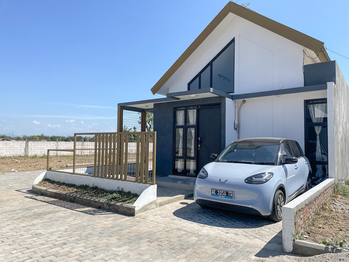 A modern house features a minimalist design with a peaked roof and large windows. A small car is parked in front on a paved driveway, with a gated entrance and a small outdoor area visible beside it. The setting is open and spacious with a clear blue sky.
