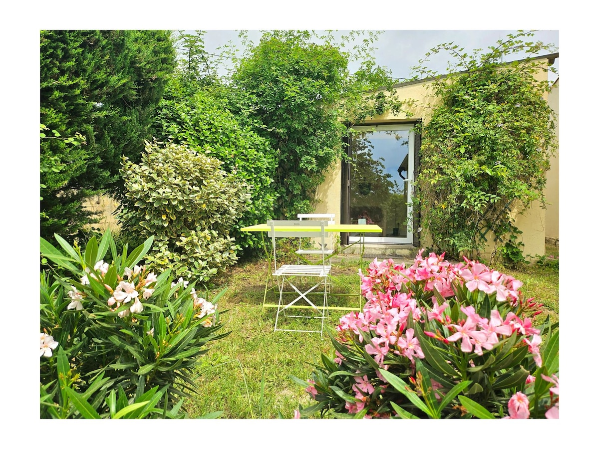 A private garden area is visible, featuring a small table and two chairs positioned amid vibrant flowers, including pink blooms. Lush greenery surrounds the space, with a glass door reflecting the garden's natural charm.