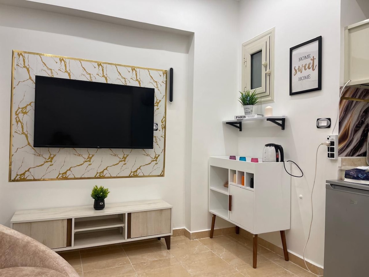 A modern living area features a large flat-screen television mounted against a stylish marble-patterned wall. Below, a light-colored media console holds decorative items. A white cabinet with open shelving and a kettle is positioned nearby, complemented by a small potted plant adding a touch of greenery.