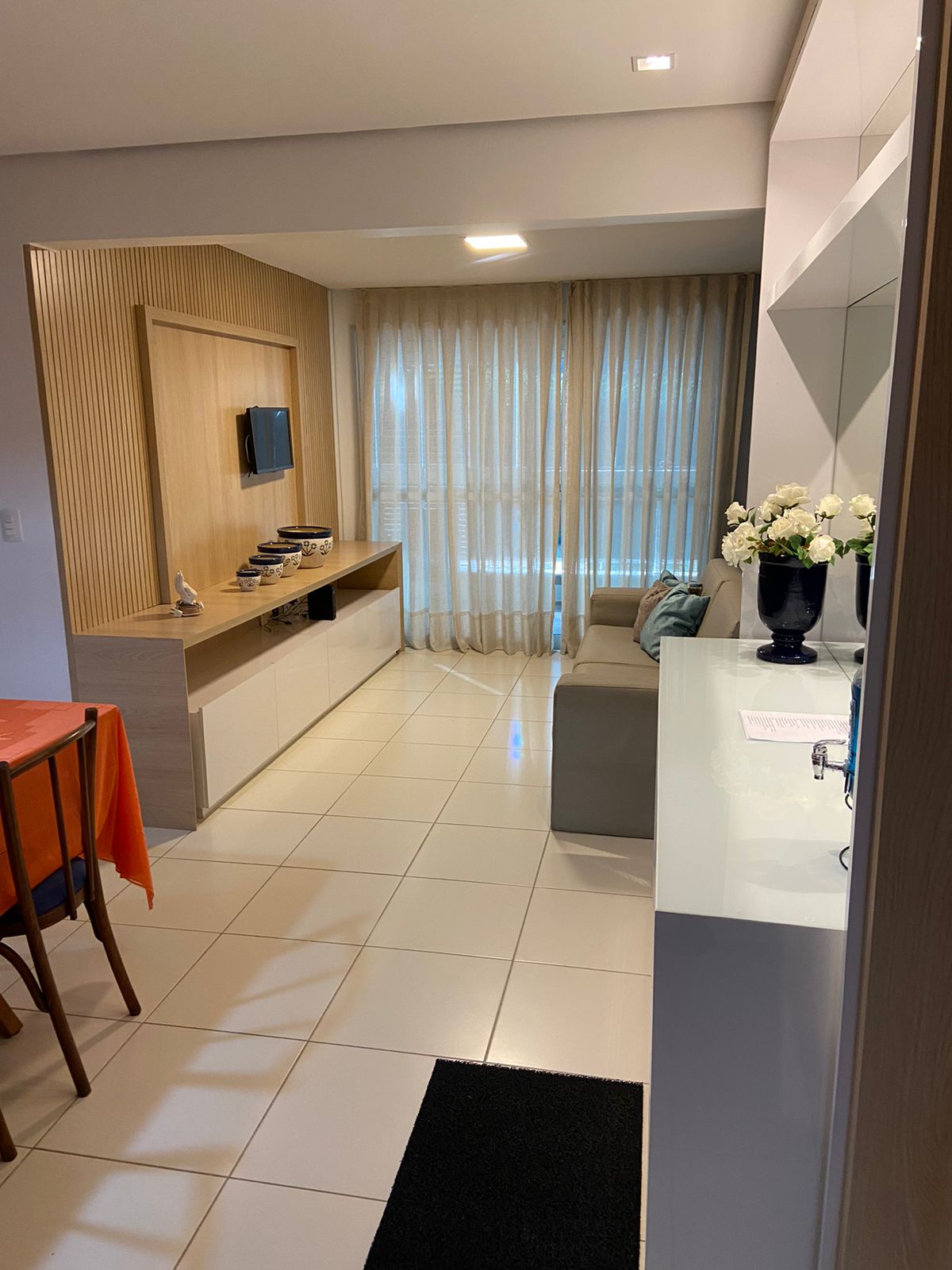 The living area features a light-filled space with large windows covered by sheer curtains. A modern sofa is positioned near a wall-mounted television, while a wooden console and a dining table with chairs are visible in the open layout.
