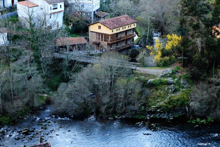 A scenic view of a riverside house surrounded by trees and natural landscape. The building features a warm-colored exterior and a large deck, overlooking the flowing water. The serene environment is complemented by the distant greenery and a bridge connecting the area.