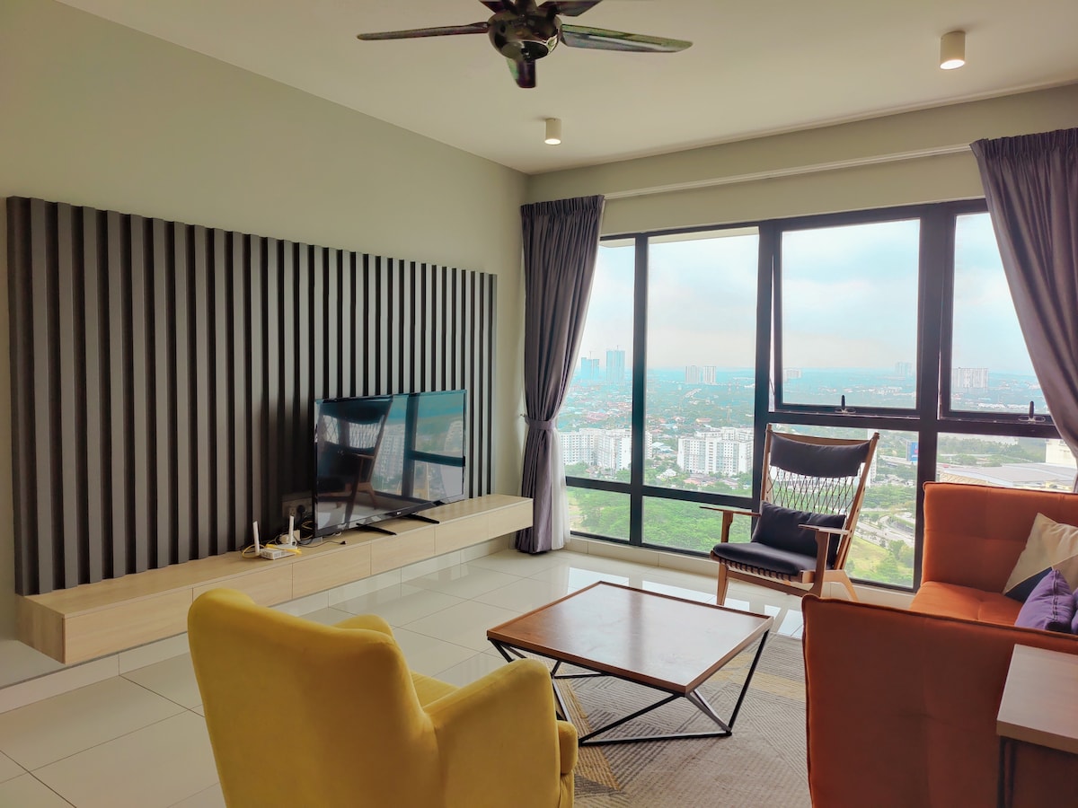 A modern living area is presented with large windows offering city views. A comfortable sofa in an orange hue and a stylish yellow armchair are arranged around a wooden coffee table. A flat-screen TV is mounted on a wooden accent wall, while soft, natural light filters through the curtains.