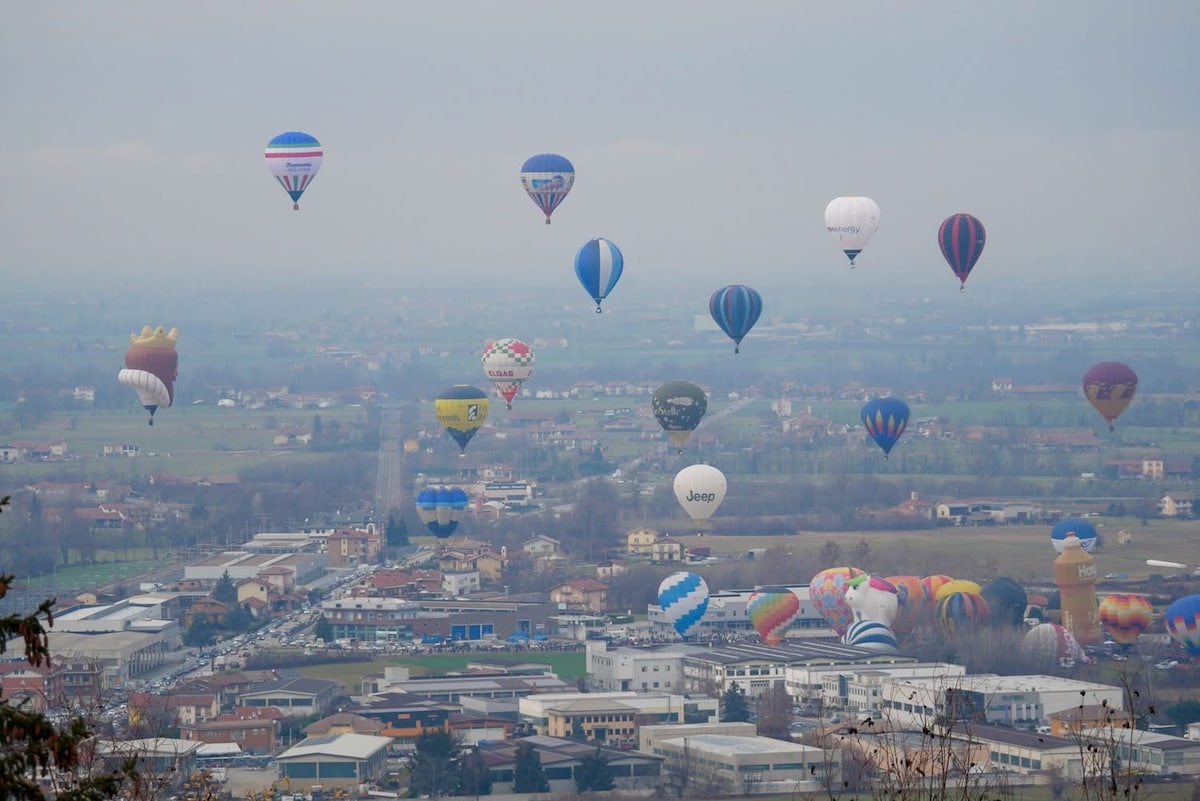 A panoramic view showcases numerous hot air balloons ascending into a cloudy sky, each displaying various colors and patterns. Below, a patchwork of fields and buildings is visible, contributing to a serene landscape that invites exploration and connection with nature.