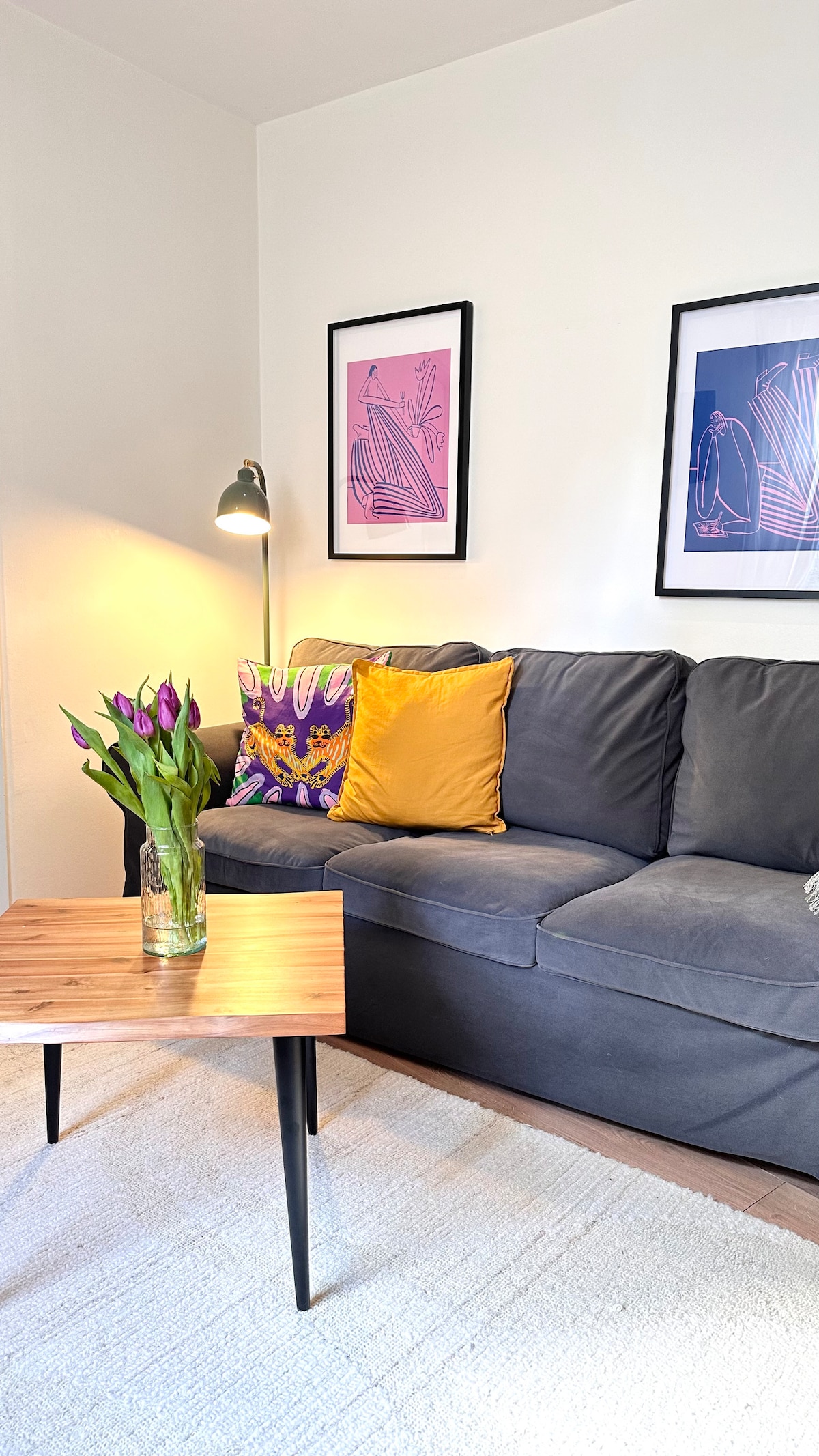 A comfortable couch is adorned with decorative pillows in vibrant colors, alongside a small wooden coffee table. A bouquet of purple tulips sits in a clear vase, while two framed artworks add a touch of elegance to the walls.