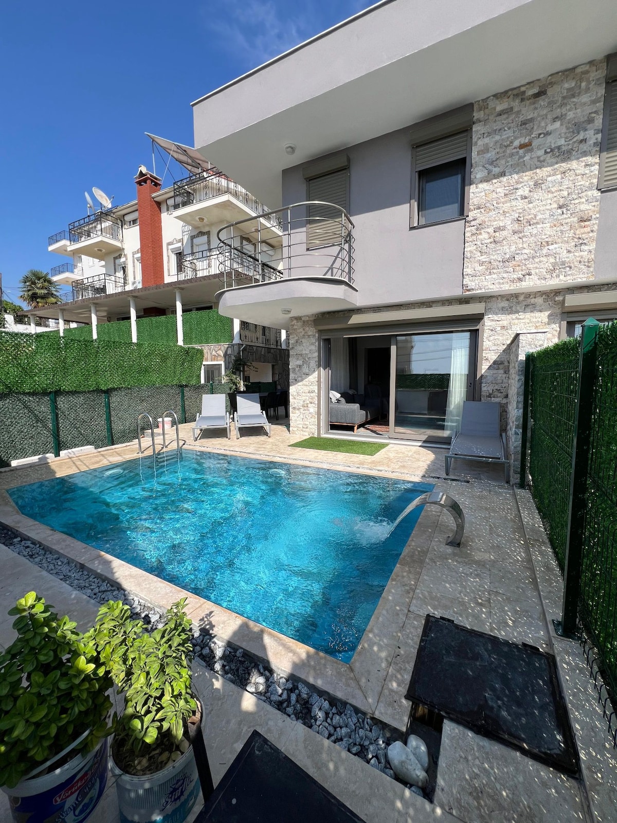 A private swimming pool is set within a stone terrace, complemented by two lounge chairs. The villa's glass doors offer views into the bright living area, while vibrant greenery provides privacy. Modern architecture features clean lines and a balcony above the pool area.