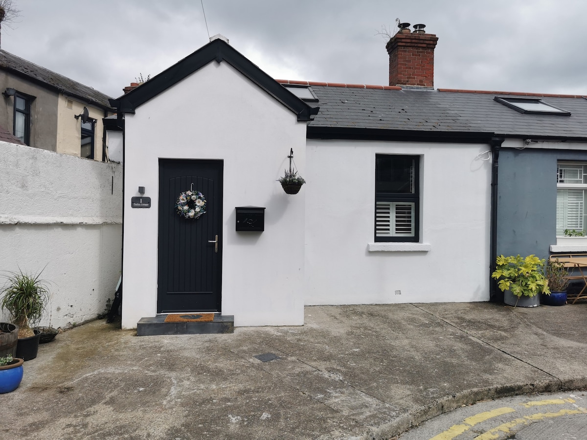 A charming cottage exterior is showcased, featuring whitewashed walls and a dark front door. A wreath adorns the door, while potted plants are visible on the ground. Windows with white shutters provide a subtle contrast, complementing the rustic feel of the setting.