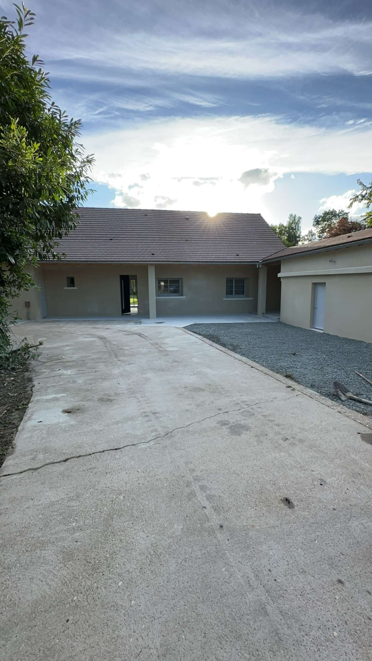 An exterior view of a recently renovated house is presented, featuring a spacious driveway with a mix of concrete and gravel. Light from the setting sun casts a warm glow on the façade, highlighting the structure's simple design and surrounding greenery.