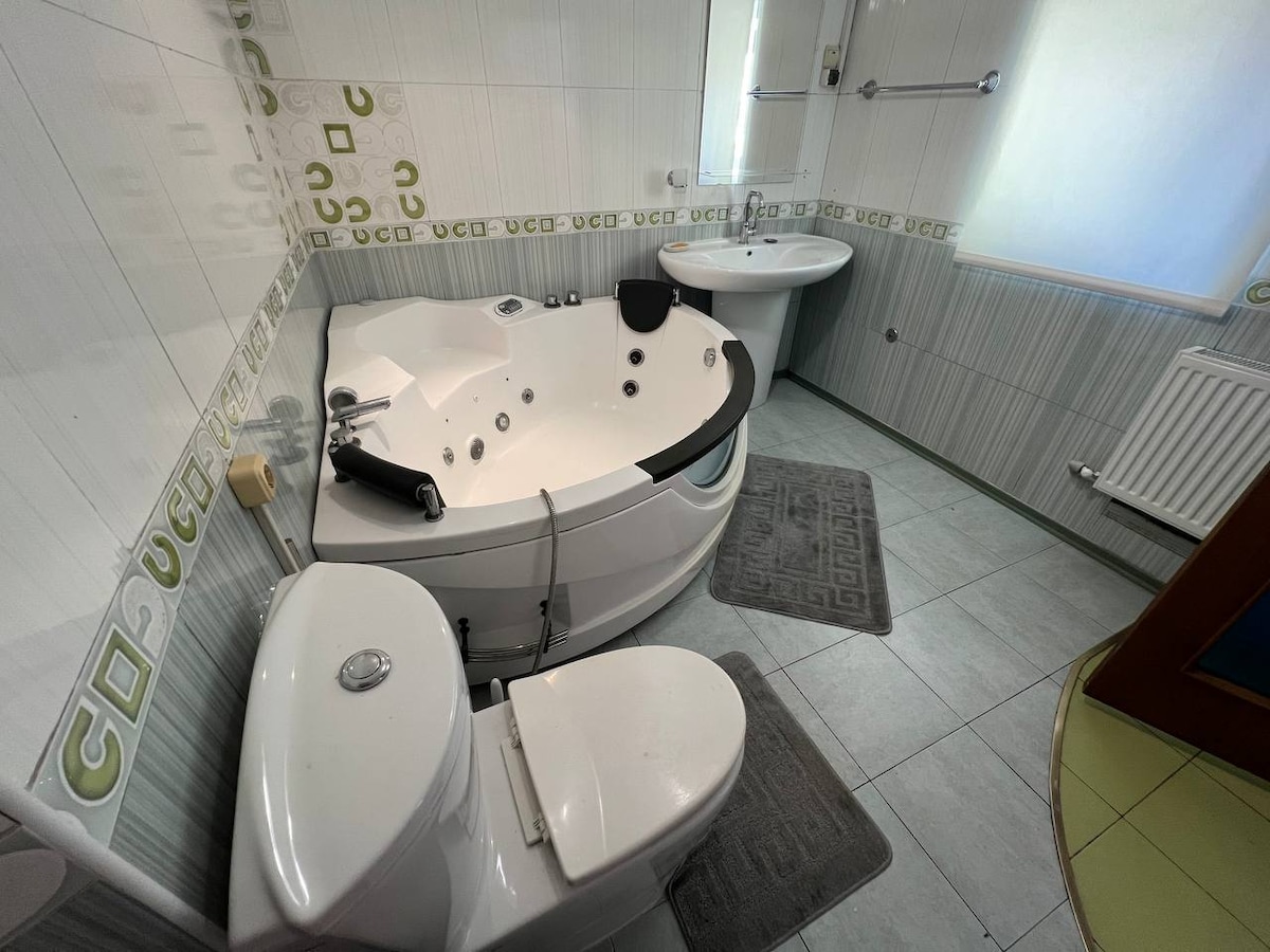 The bathroom contains a modern whirlpool bathtub, paired with a sleek toilet and a stylish sink. Soft, neutral tones are complemented by subtle decorative accents on the walls. Natural light enters through a nearby window, contributing to a bright and clean atmosphere.