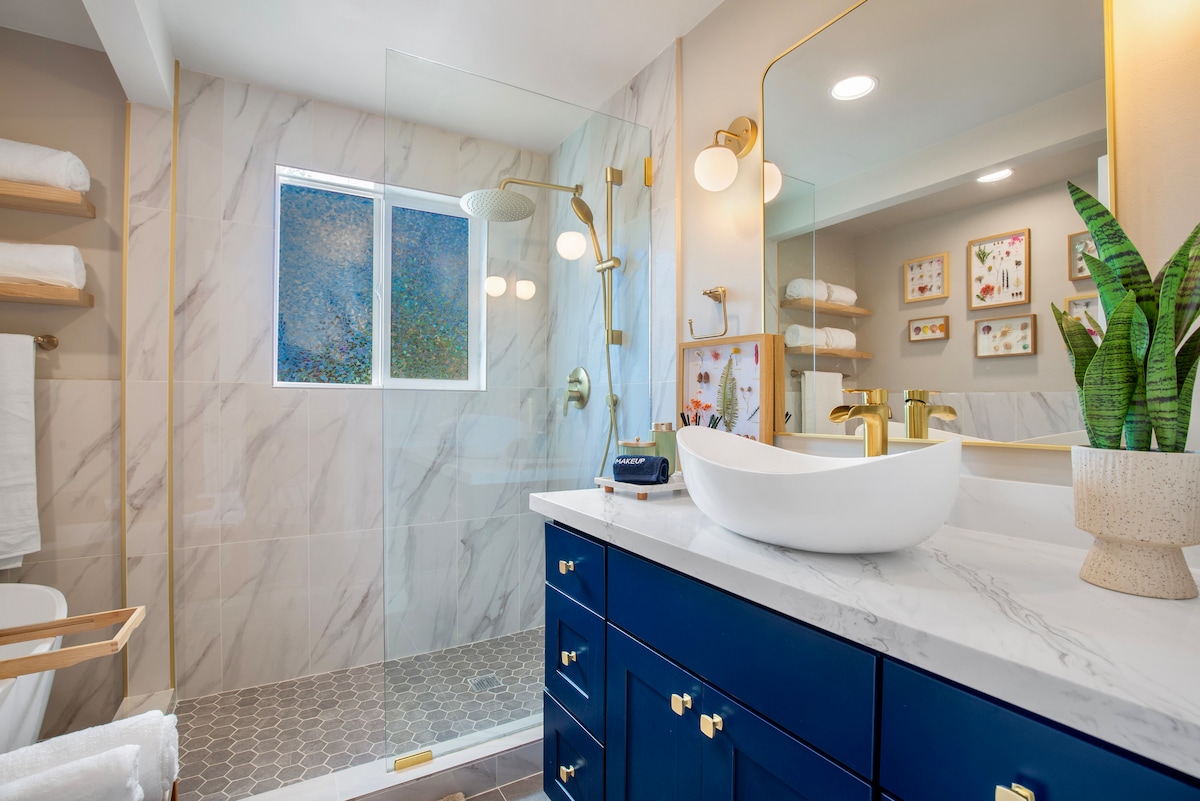 The bathroom features a glass-enclosed shower with a rainfall showerhead, complemented by marble walls. A floating vanity with a modern vessel sink is paired with navy cabinetry. Shelves hold neatly arranged towels, and artwork adorns the walls, adding a touch of color.