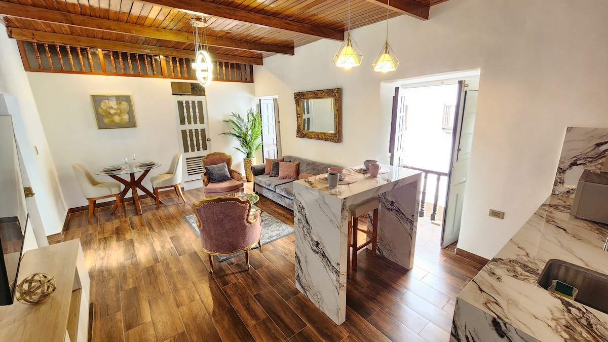 A modern living space features a comfortable sofa and a dining table set for two, illuminated by stylish pendant lights. Large windows provide natural light, while the decorative mirror adds depth. The kitchen area presents elegant marble countertops, enhancing the contemporary design.