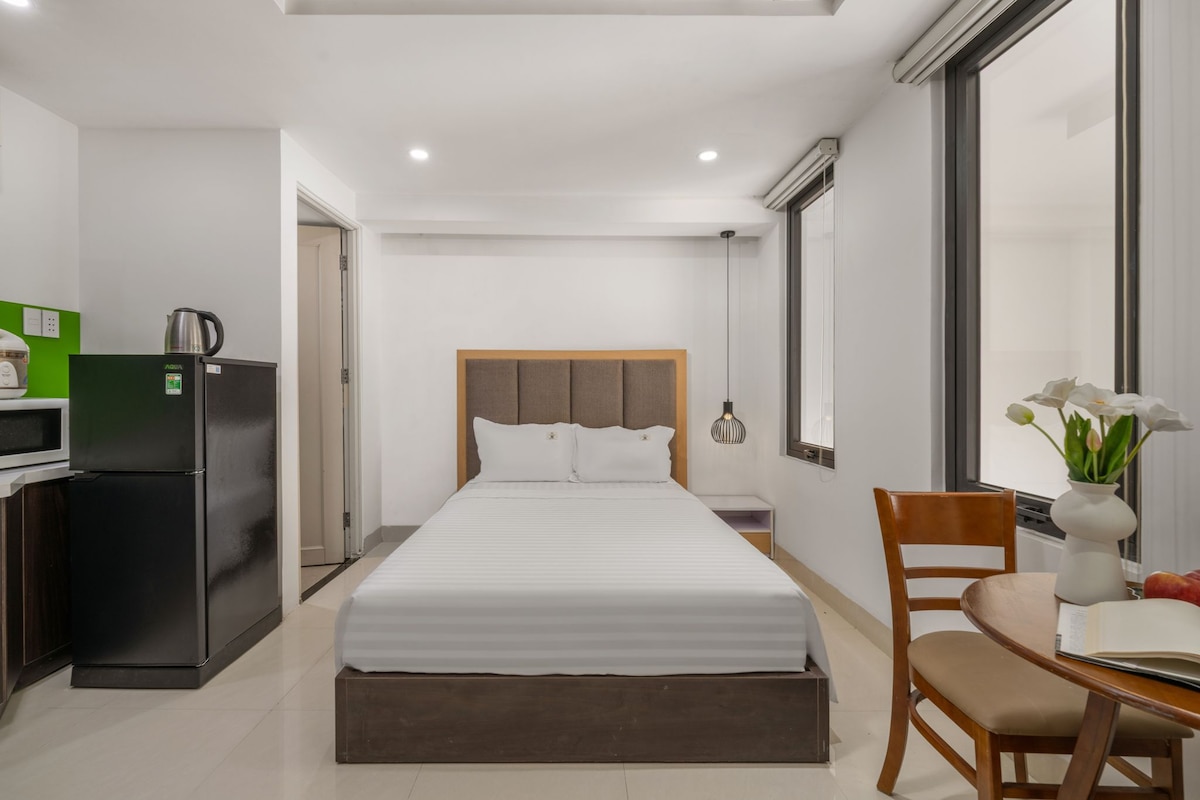 A bright and spacious room features a comfortable bed with crisp white linens. A small dining table is set near a window, and a kitchenette is equipped with a sink and microwave. A modern aesthetic is complemented by simple decor and natural light.